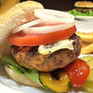 a pork burger on a bun dressed with roasted peppers, feta cheese, tomato, dill pickle slices onion and lettuce all on a plate