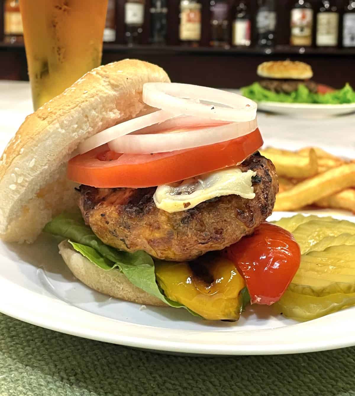 pork burger on a bun with roasted peppers cheese lettuce tomato onion on a plate with pickle slices and french fries