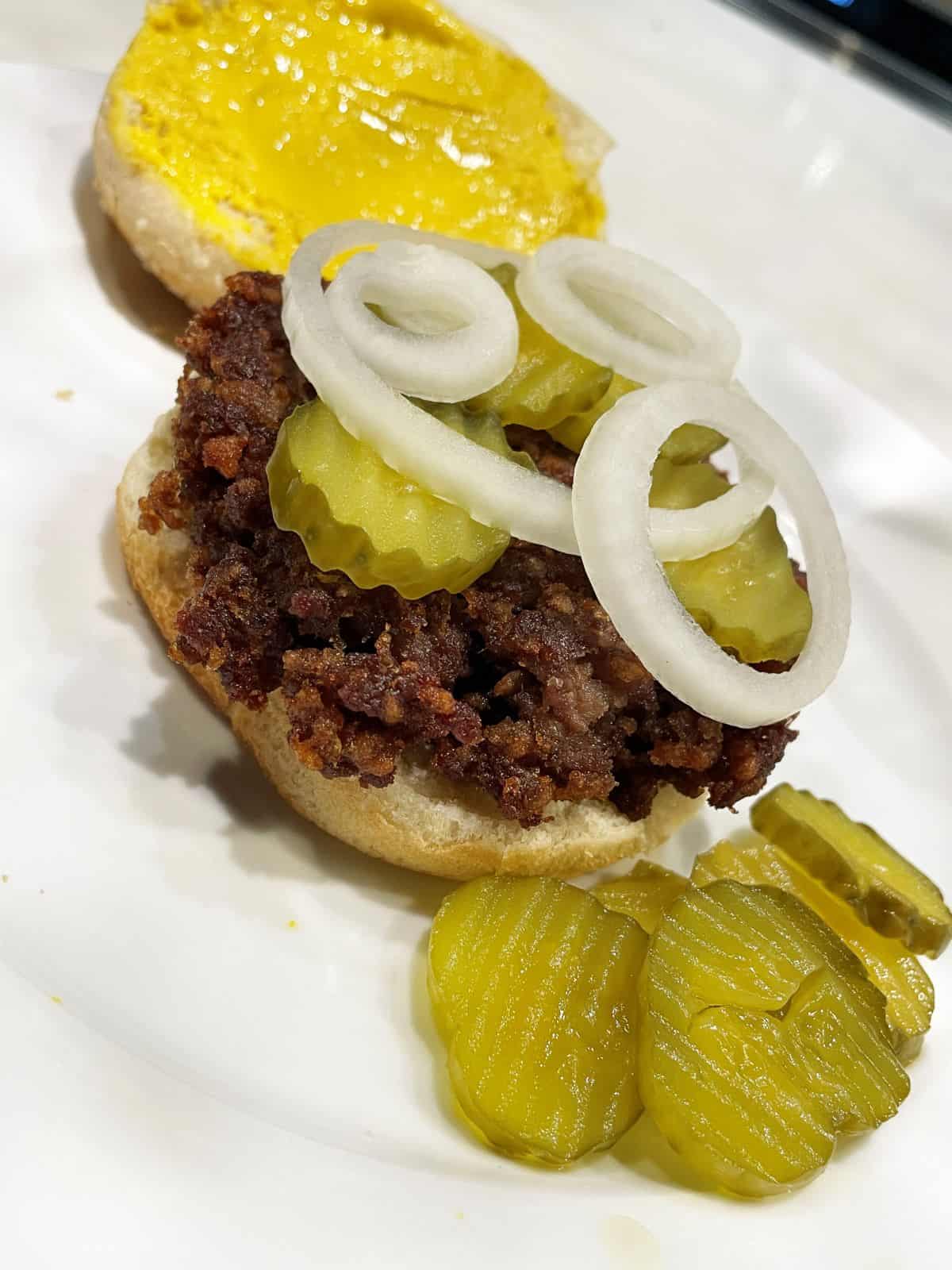 slugburger on a bun with pickles and onion with the top bun in the back has mustard on it and more pickles on the side