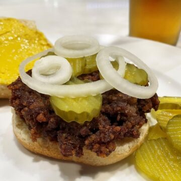 slugburger on a bun with pickles and onion with the top bun in the back has mustard on it and more pickles on the side and a glass of beer next to the plate