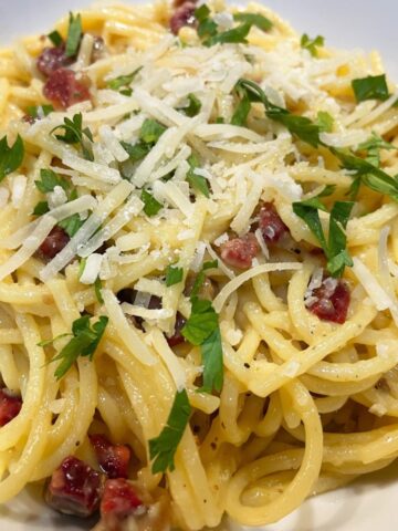 spaghetti carbonara in a white bowl garnished with chopped parsley and shredded romano cheese