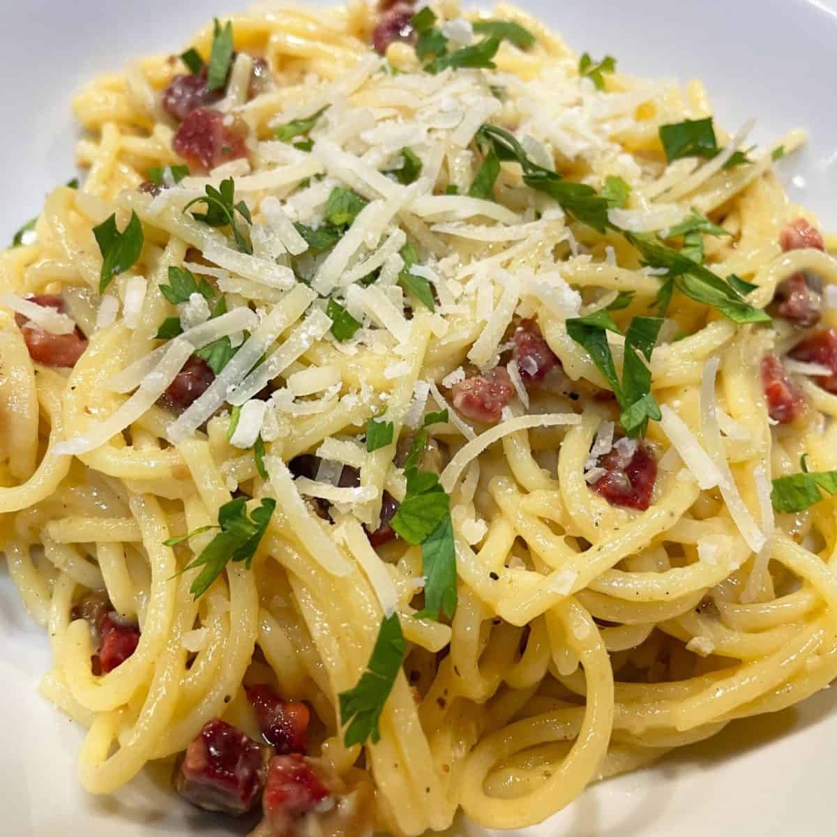 spaghetti carbonara in a white bowl garnished with chopped parsley and shredded romano cheese